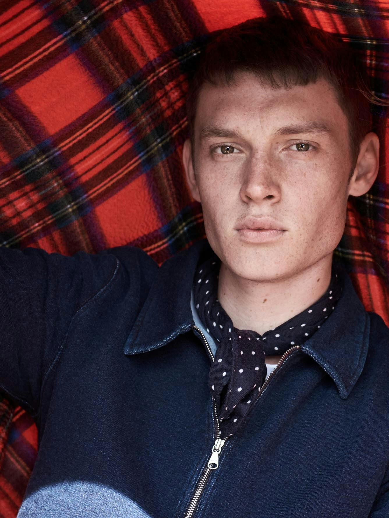 Male Model Wearing a navy neckerchief with white spots and a navy sports code holding a red tartan blanket behind him