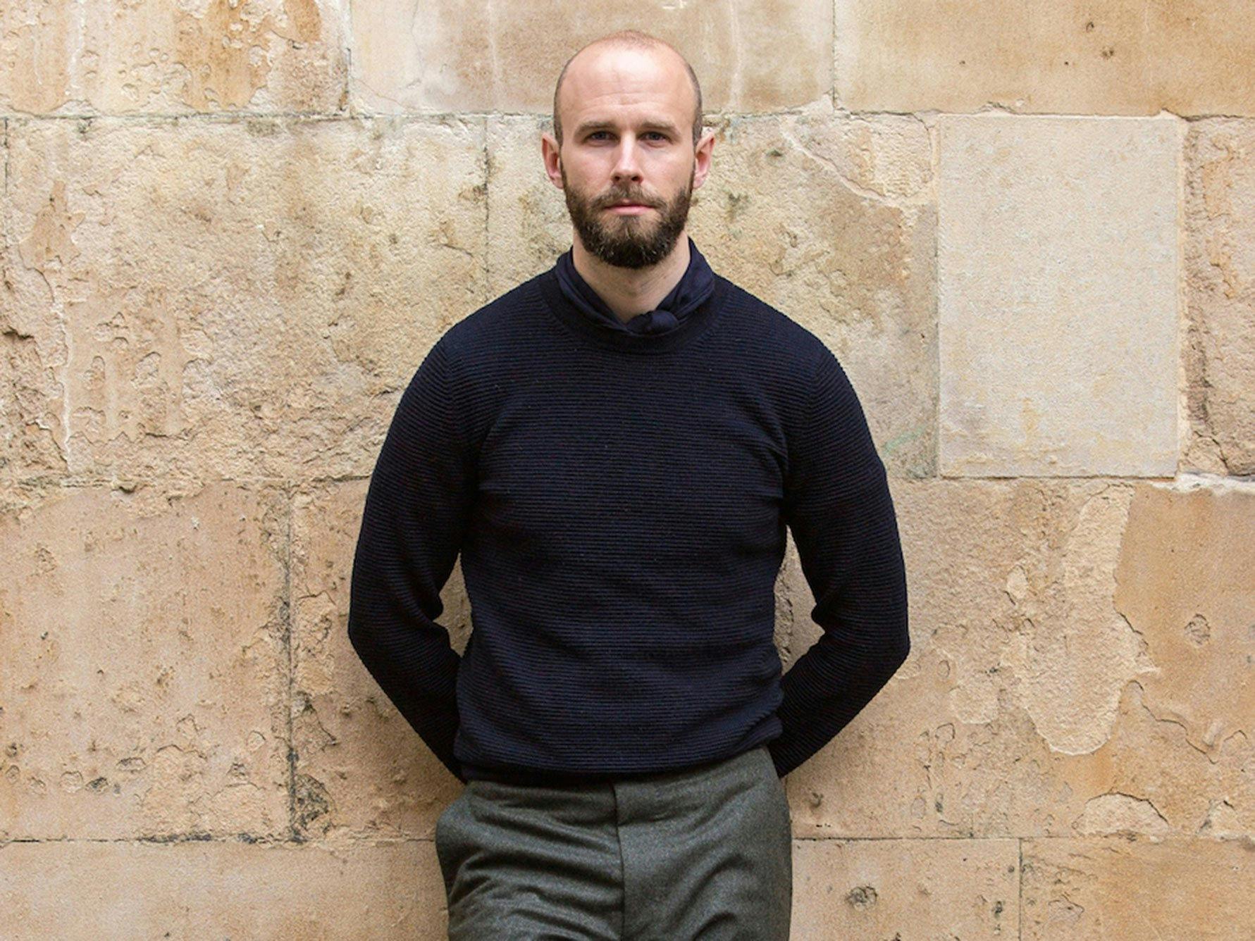 Simon Crompton leans against a brick wall wearing a navy jumper with navy cashmere square neckerchief and grey slacks